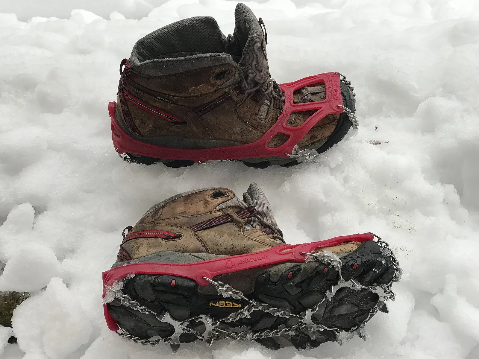 雪の上に置かれた2足の登山靴に、赤いバンドのチェーンスパイクが装着されている