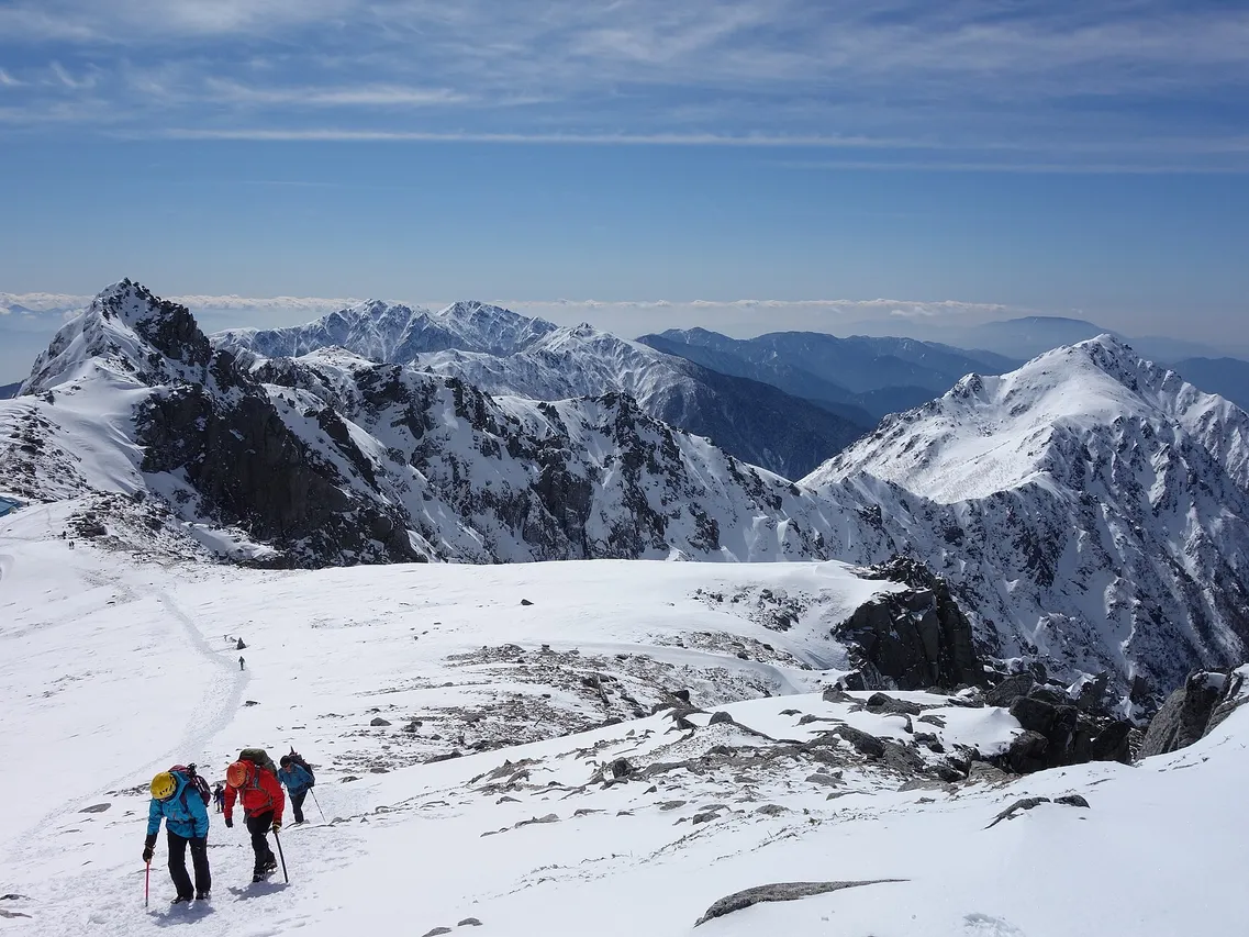 中央アルプスの雪に覆われた稜線を、登山者たちが列になって登っていく様子。背後に雪をかぶった山並みと青空が広がる