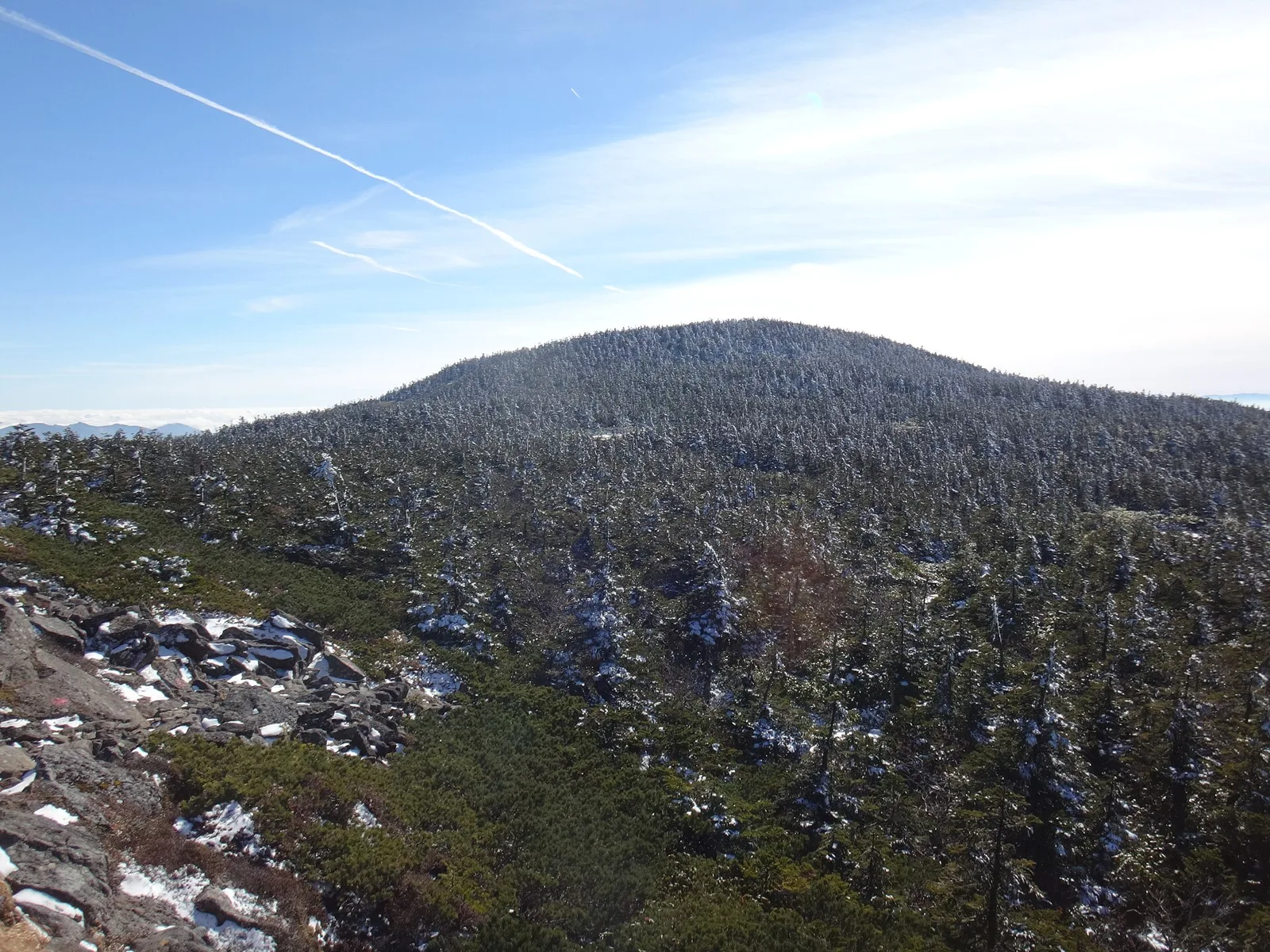 晴れた空の下、樹木に雪や霧氷が付いて白くなった西吾妻山の山頂部と、手前の雪の残る岩場