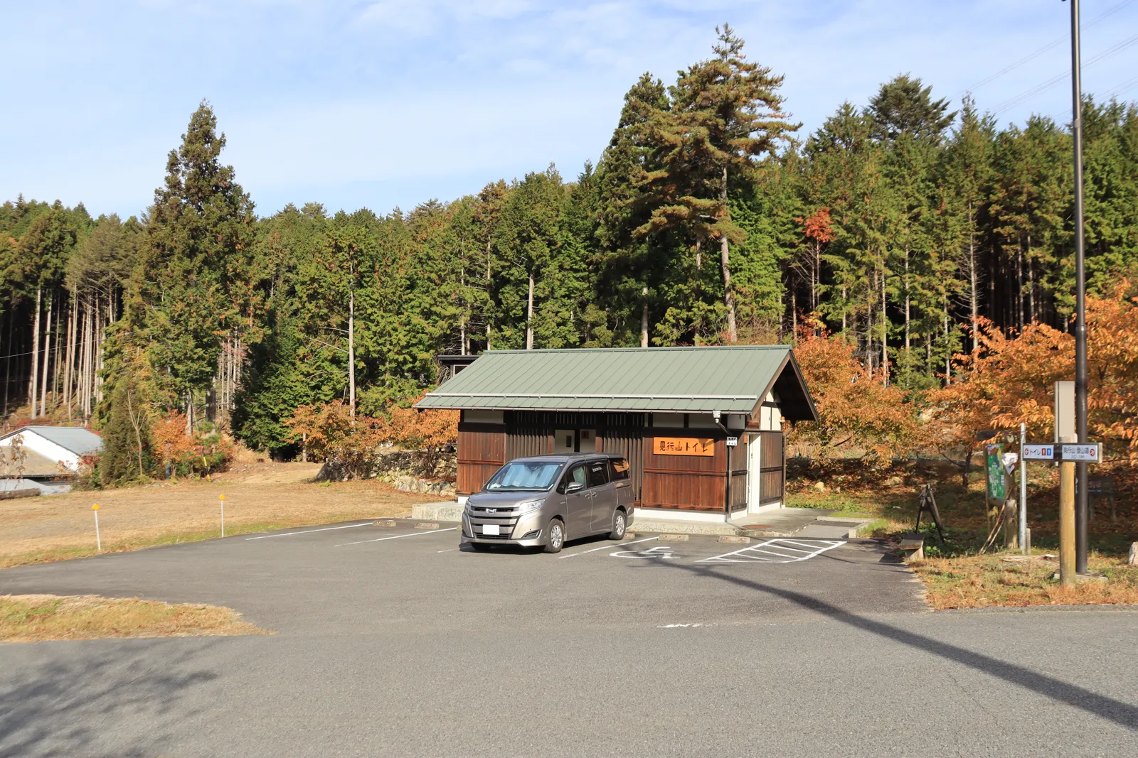 杉林と色づいた木々に囲まれた登山口の舗装された駐車場に、車が1台とまり、奥に小さなトイレの建物がある