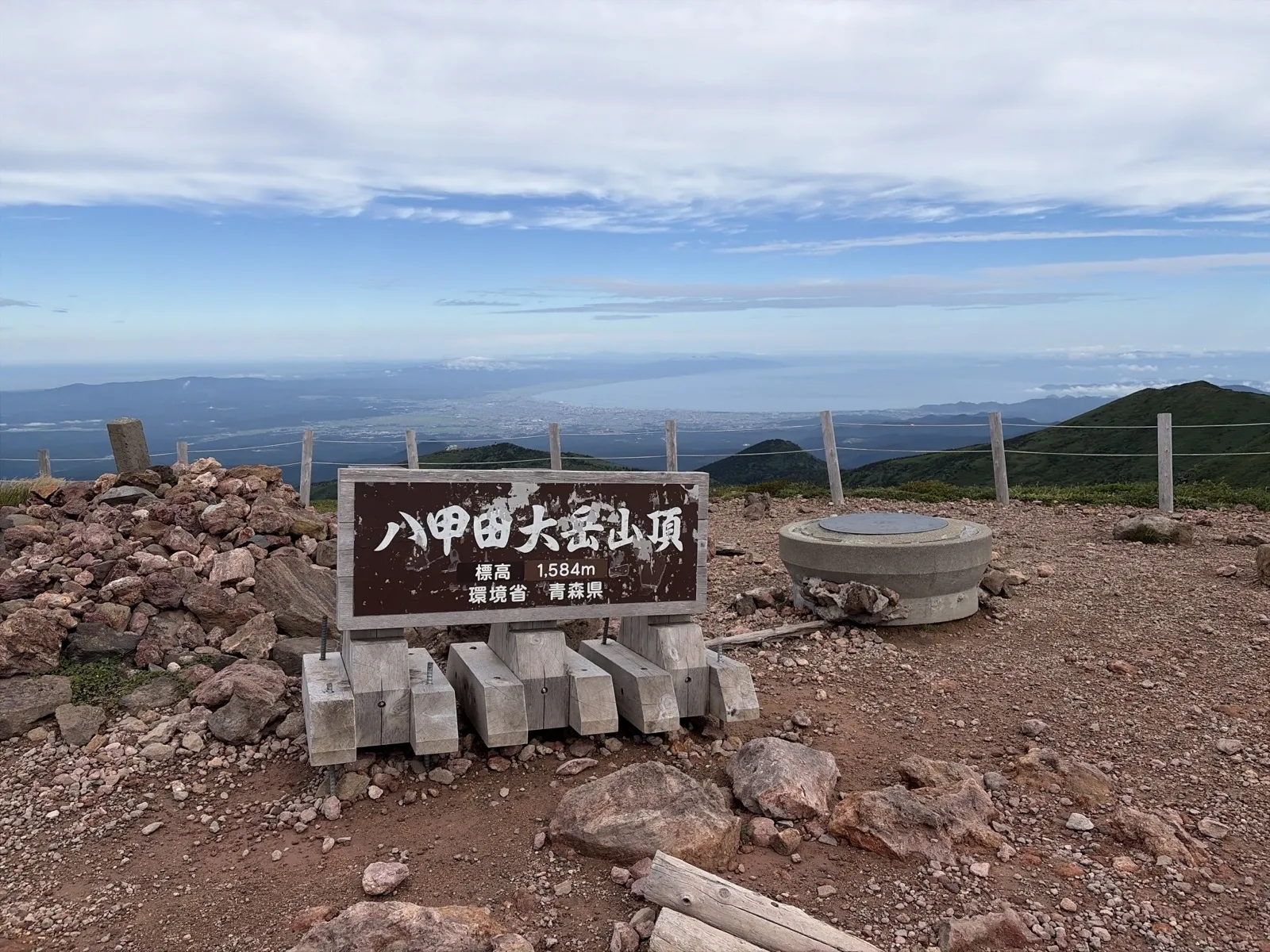 赤茶色の土と岩の山頂に立つ「八甲田大岳山頂 標高1,584m 環境省 青森県」の標識。奥には海と海沿いの街並みが広がり、空には雲がかかっている