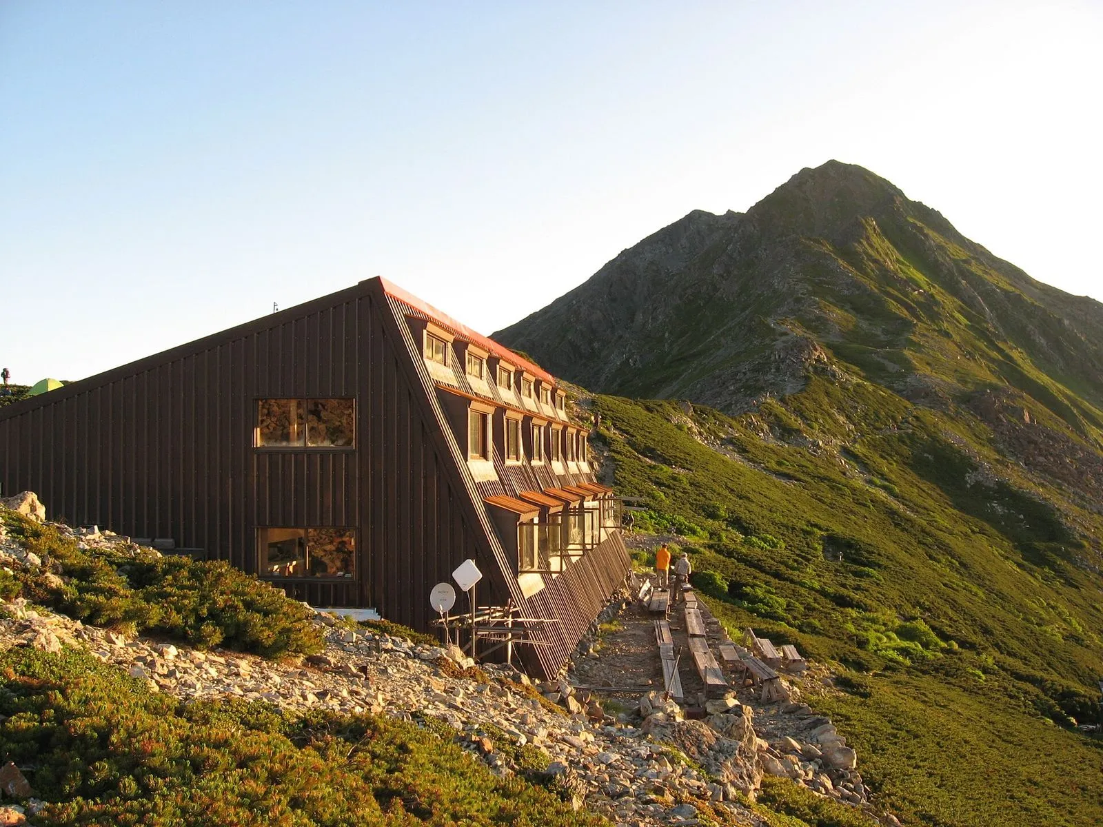 A mountain hut in the evening light below a rocky peak in the Southern Alps, with hikers walking on the boardwalk beside it