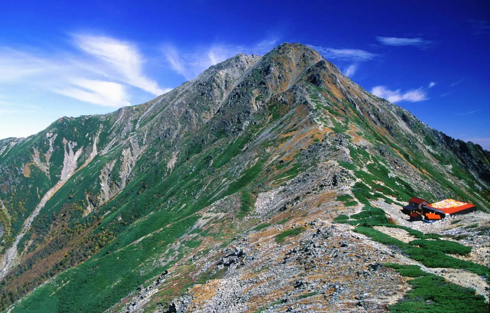 高山の稜線上の鞍部に建つ山小屋と、その奥にそびえる岩の山