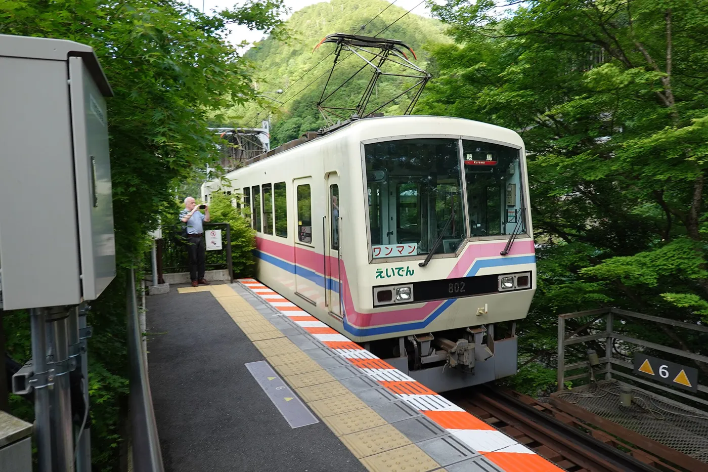 A white two-car Eizan Railway train with red and blue stripes at the forested Kibuneguchi Station on the Kurama Line