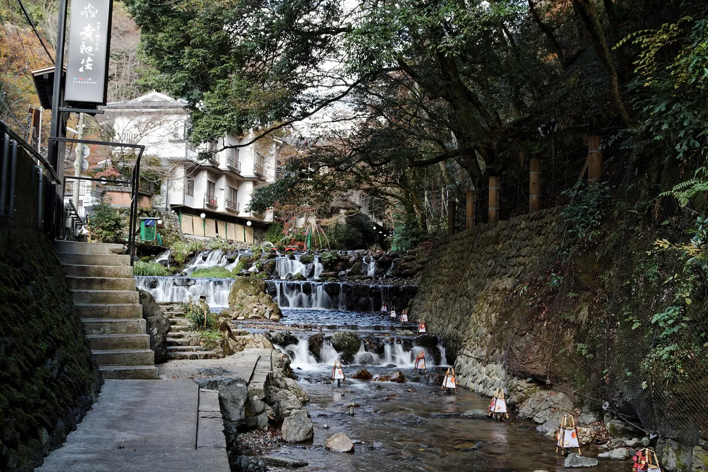The narrow Kibune River flowing over rocks beside the village road, with wooden restaurant buildings and autumn trees along the bank