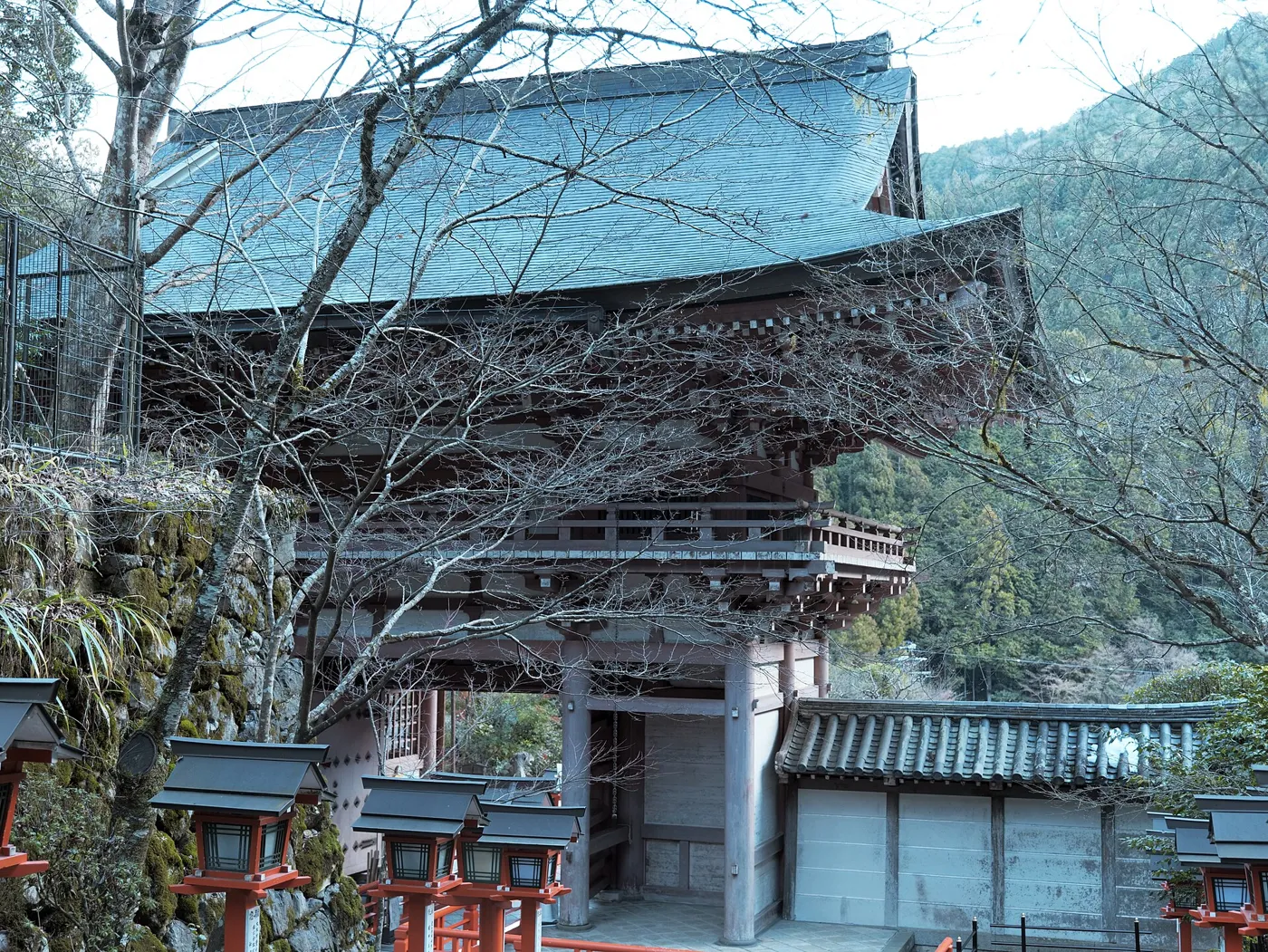The red-painted two-storey Nio-mon gate of Kurama-dera, seen from inside the temple grounds, with forested slopes behind
