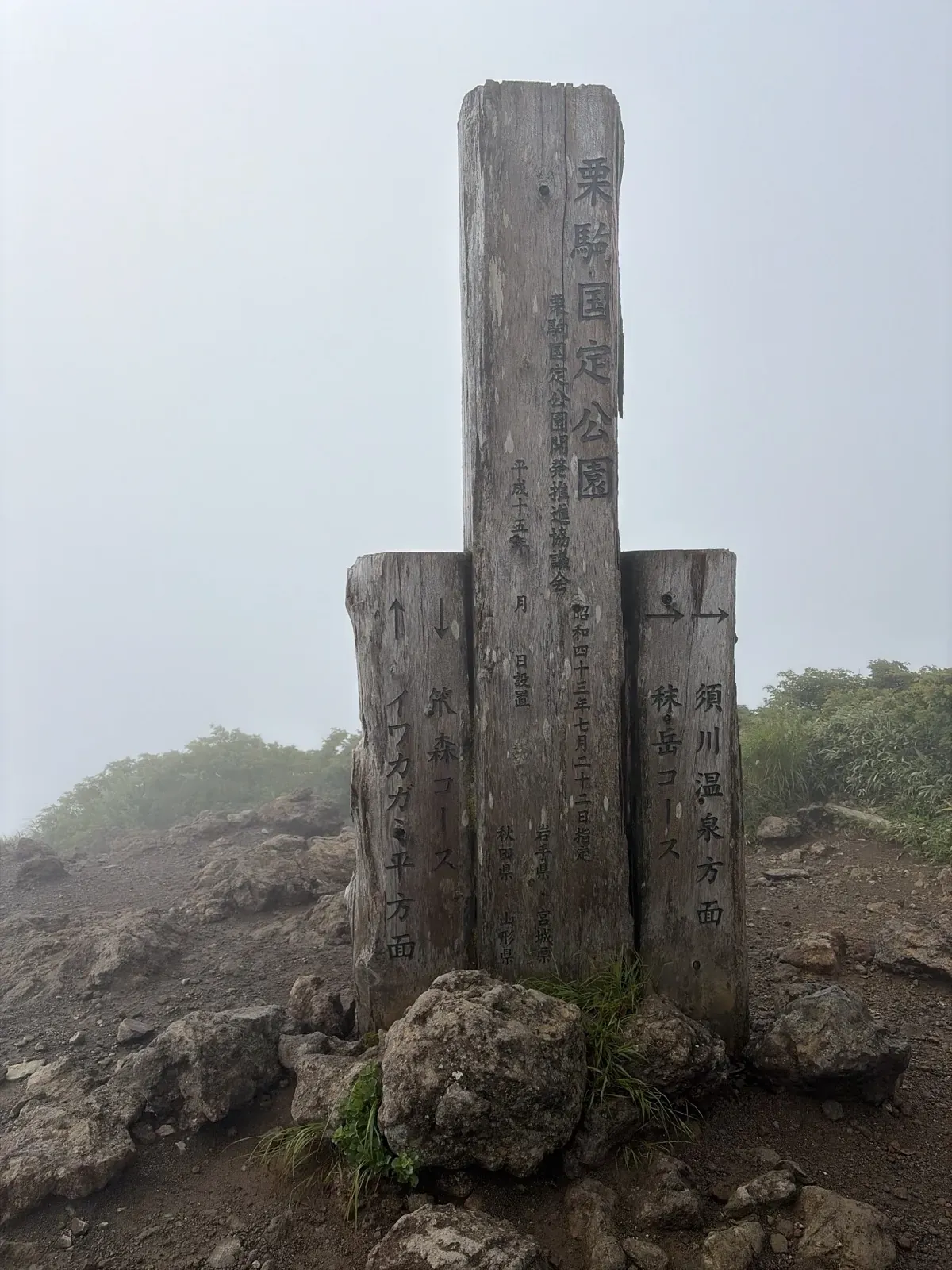 霧の栗駒山の山頂に立つ「栗駒国定公園」の木の標柱。左右の柱にイワカガミ平方面・笊森コース、須川温泉方面・秣岳コースの文字