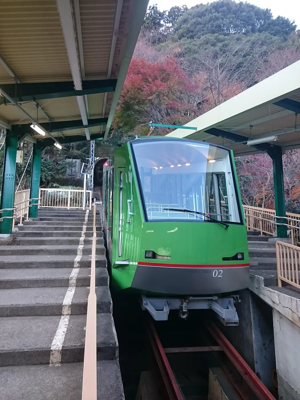 A green Oyama Cable Car car stopped at a station platform with stepped walkways and autumn trees around it