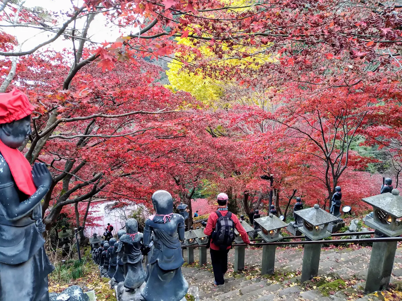 Stone steps at Oyama-dera Temple covered by bright red and yellow maple leaves, with small Buddhist statues along the side and a hiker walking down