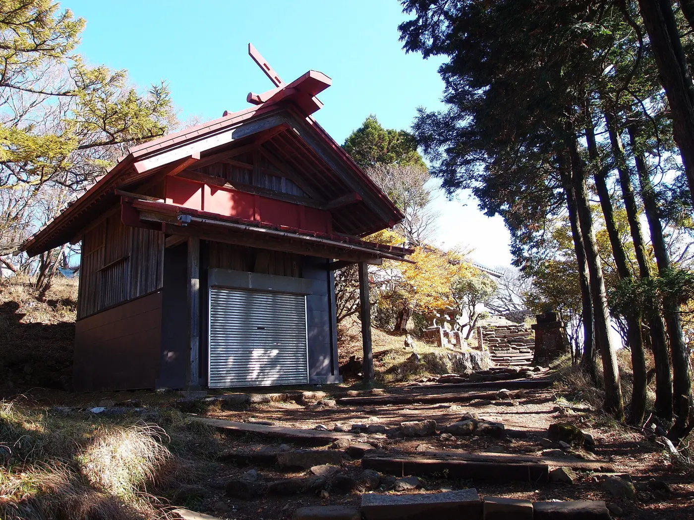 A red-painted wooden shrine building on the summit of Mount Oyama beside a stone-and-earth path, with tall trees and a blue sky