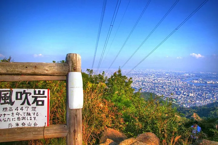 The wooden sign for Kazafuki-iwa beside a rocky viewpoint, with the city and Osaka Bay spread out below under a blue sky