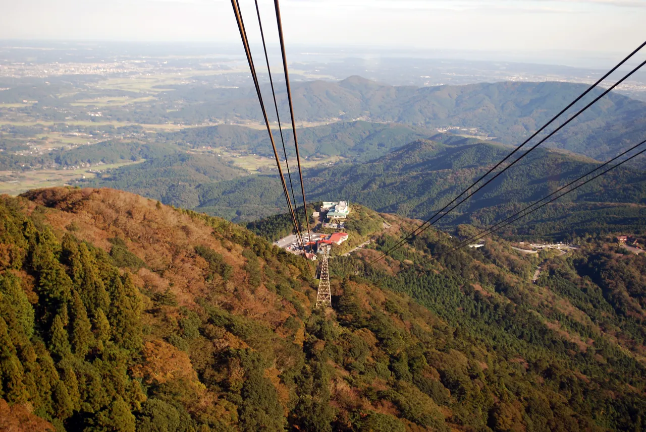 从高处俯瞰筑波山空中缆车的缆索和支柱，山坡上的树林开始变成红褐色，远处是平原和低矮的山丘