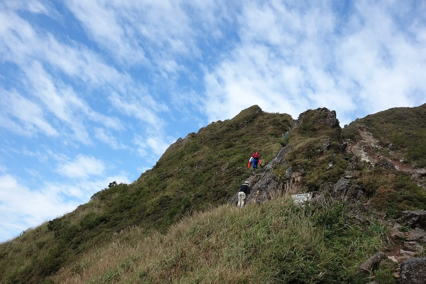 A hiker using a chain on a steep rocky ridge on the way to the West Peak of Mount Yufu, with grass and blue sky around