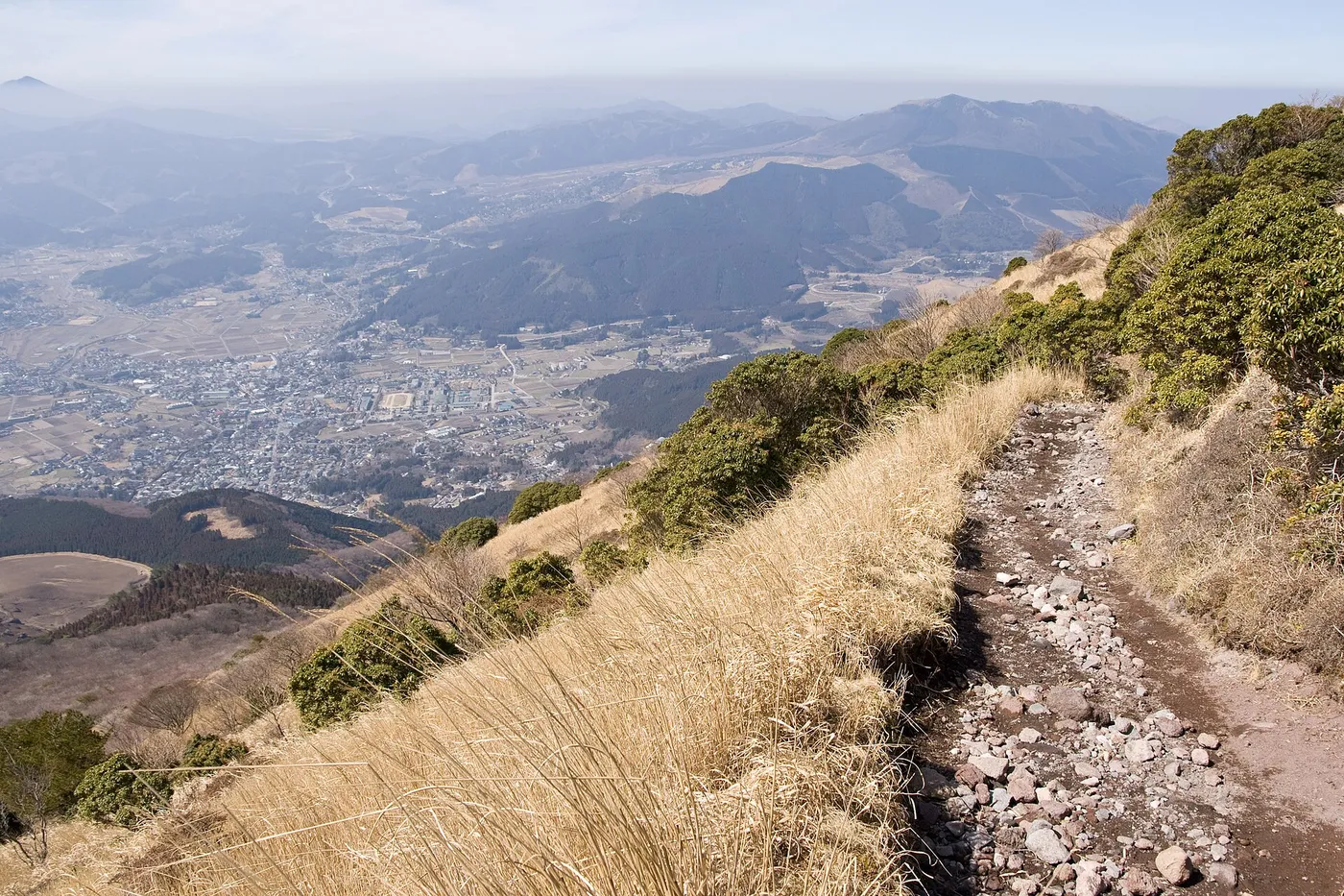 A rocky hiking trail climbing through dry grass and low shrubs on the slope of Mount Yufu, with the town of Yufuin spread across the basin far below