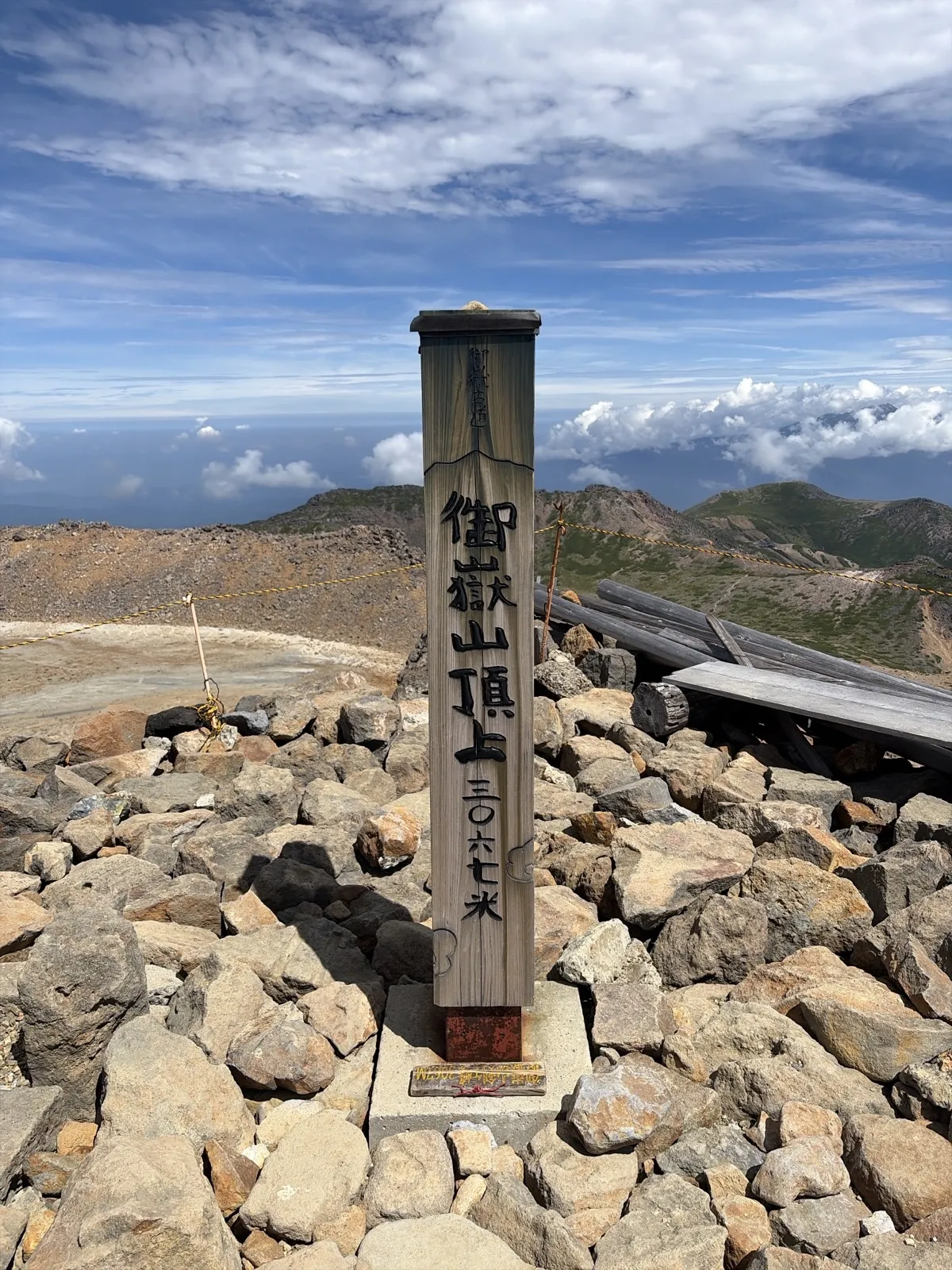 岩が積み重なる山頂に立つ木の標柱。「御嶽山頂上 三〇六七米」と書かれ、奥には青空と雲が広がっている