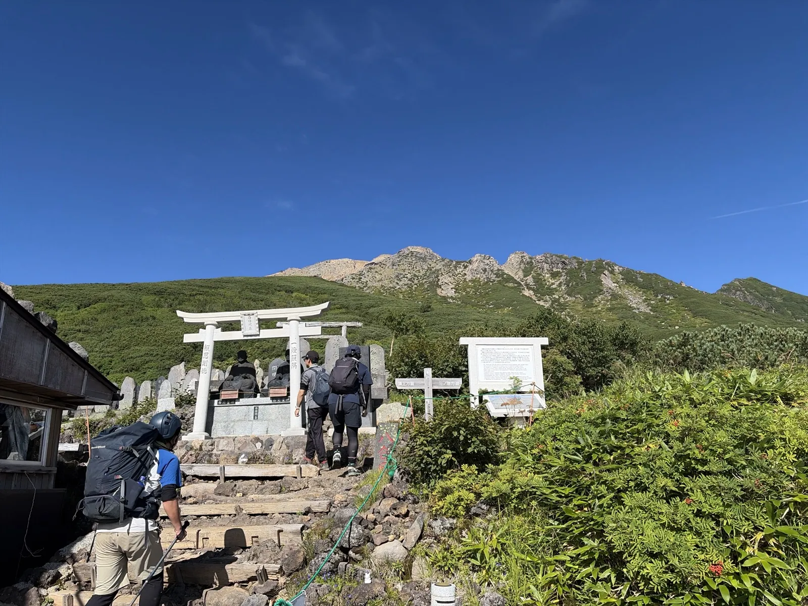 白い鳥居と石像が並ぶ場所に登山者が集まっている。奥には青空の下に岩の山頂部が見える