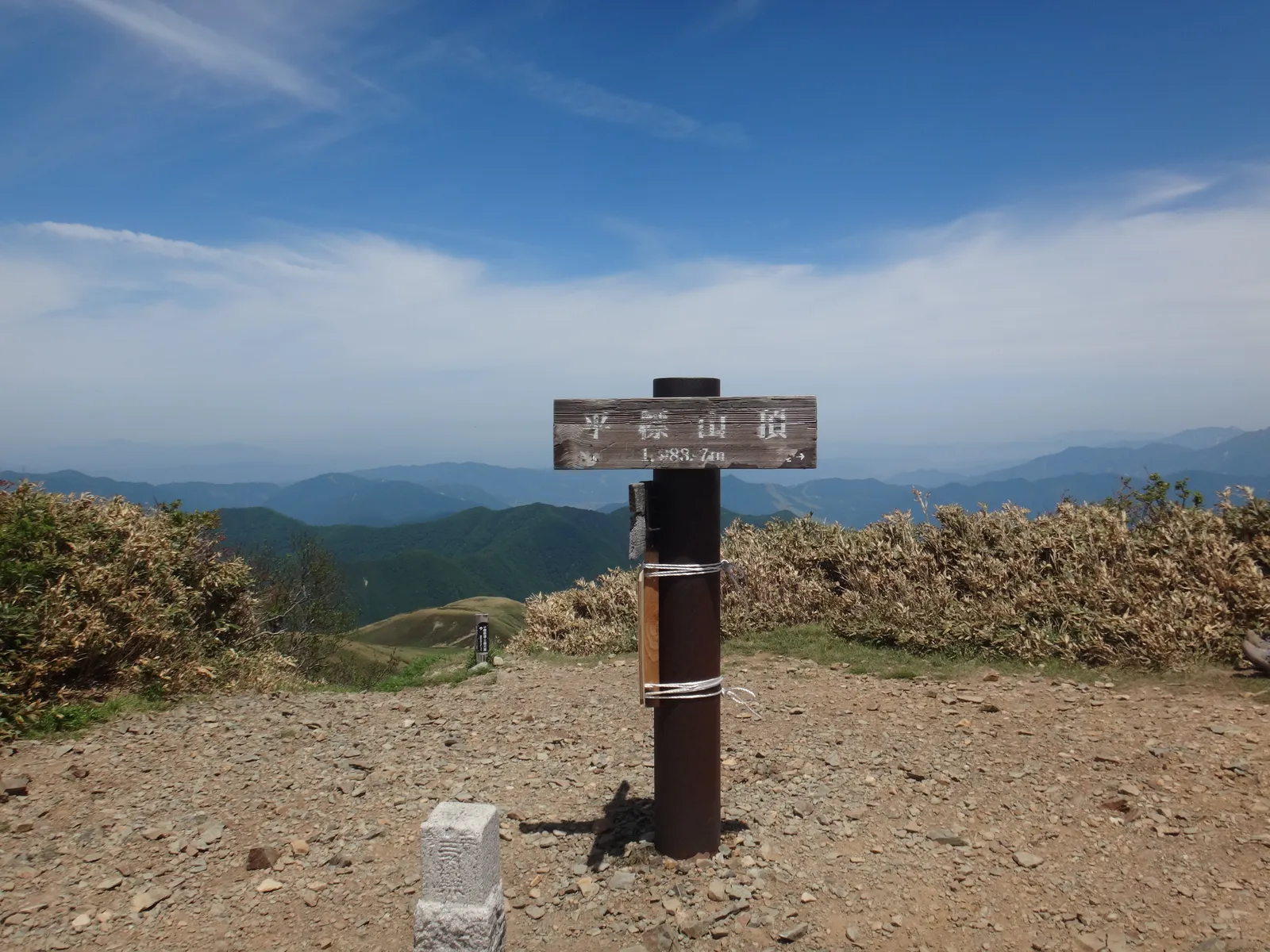青空の下、平らに開けた平標山の山頂に立つ山頂標柱と、遠くに連なる山並み