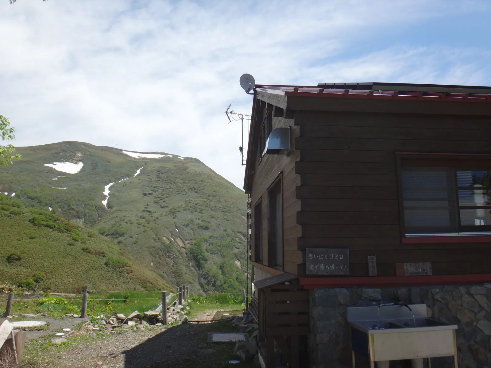 青空の下に建つ平標山の家の建物と、その奥に見える残雪の山の斜面