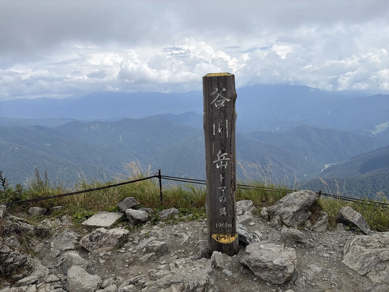 ロープで囲まれたトマノ耳の山頂に立つ木の標柱。「谷川岳 トマの耳 1963m」と書かれ、奥には緑の山並みと雲が広がっている